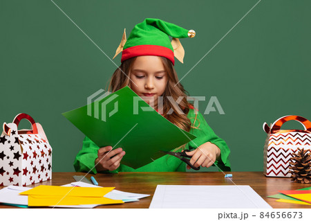 Little cute girl dressed like funny gnome or elf sitting at table and making paper Christmas decorations isolated over green background. 84656673