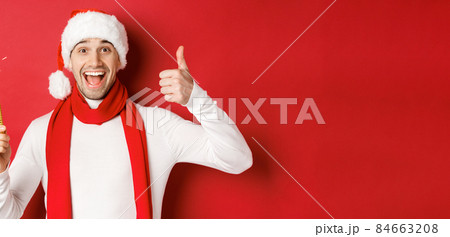 Concept of christmas, winter holidays and celebration. Handsome man celebrating new year and having fun, holding sparkler and showing thumb-up, standing over red background 84663208