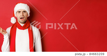 Concept of christmas, winter holidays and celebration. Portrait of confused man in santa hat and scarf, frowning and looking perplexed, standing over red background 84663209