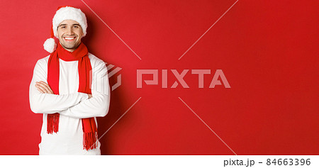 Handsome caucasian guy with bristle, wearing santa hat, scarf and white sweater, cross arms on chest and smiling happy, standing against red background 84663396