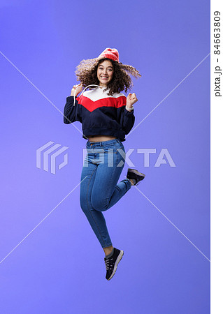 Delighted carefree and happy young woman with curly hair in warm beanie jumping joyfully having fun raising hands and smiling broadly posing over blue background in outdoor clothes 84663809