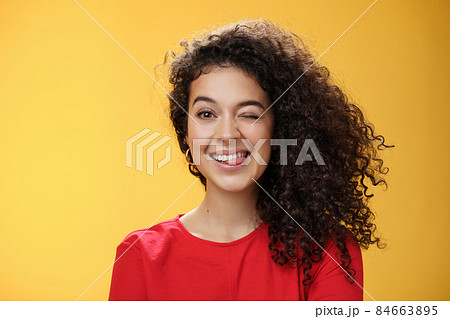 Portrait of funny and cool sister with curly hair winking playfully having fun and foolind around showing tongue as playing with siblings adoring spend time with chidren over yellow background 84663895