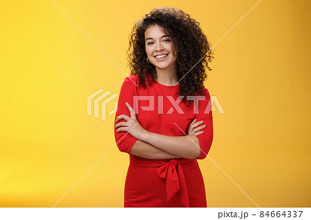 Confident charismatic young curly-haired woman in casual red dress holding hands crossed over chest in self-assured satisfied expression, smiling delighted posing over yellow background 84664337