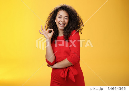 Girl feeling awesome. Friendly-looking excited and upbeat charming girlfriend with curly hair in dress winking playfully and smiling as showing okay gesture having deal and things under control 84664366