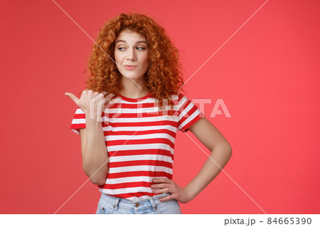 Look that stuff. Redhead curious sassy good-looking curly-haired girl summer striped t-shirt smirking gazing pointing left indicating thumb intriguing product standing red background Look that stuff. Redhead curious sassy good-looking curly-haired girl summer striped t-shirt smirking gazing pointing left indicating thumb intriguing product standing red background 84665390