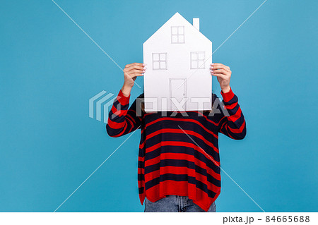 Unknown woman wearing striped casual style sweater, hiding her face behind paper house with drawn windows and door, government housing program. Indoor studio shot isolated on blue background. 84665688
