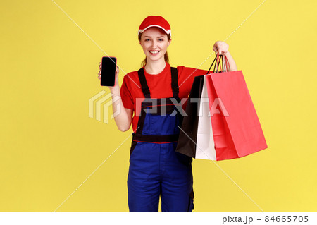 Portrait of happy positive courier woman standing with shopping bags in hands and showing smart phone with blank screen for ad, wearing overalls. Indoor studio shot isolated on yellow background. 84665705