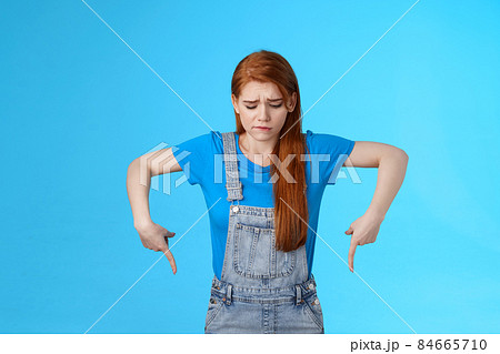 Moody upset cute redhead woman feel uneasy, frowning look pointing up jealousy and regret, cannot afford awesome new product, express sadness and sorrow, stand blue background gloomy 84665710
