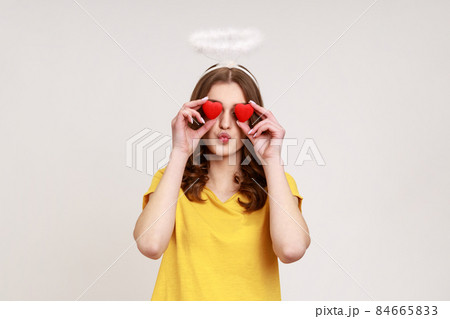 Romantic young female halo covering eyes with toy hearts as if looking with love, making kissing gesture, affection adoration in relations. Indoor studio shot isolated on gray background. Romantic young female halo covering eyes with toy hearts as if looking with love, making kissing gesture, affection adoration in relations. Indoor studio shot isolated on gray background. 84665833