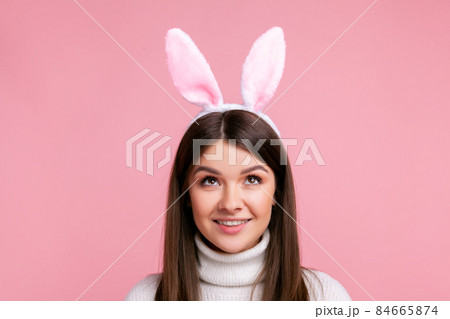 Young female with bunny ears looking up with playful attractive smile, childish carefree expression, wearing white casual style sweater. Indoor studio shot isolated on pink background. 84665874