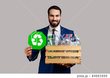 Bearded man holding box with plastic bottles and recycling green symbol, sorting his rubbish, saving ecology, wearing official style suit. Indoor studio shot isolated on gray background. Bearded man holding box with plastic bottles and recycling green symbol, sorting his rubbish, saving ecology, wearing official style suit. Indoor studio shot isolated on gray background. 84665899