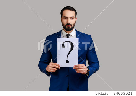 Portrait of serious bearded businessman looking at camera, holding paper with question mark, thinks about tasks, wearing official style suit. Indoor studio shot isolated on gray background. Portrait of serious bearded businessman looking at camera, holding paper with question mark, thinks about tasks, wearing official style suit. Indoor studio shot isolated on gray background. 84665921
