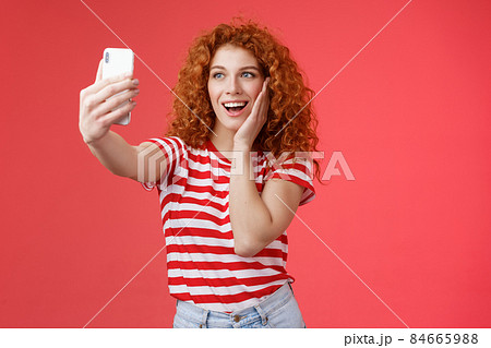 Fashionable cute redhead curly female summer t-shirt record video social media from luxury tropical resolt travel holidays abroad take selfie hold smartphone pose silly photograph red background 84665988