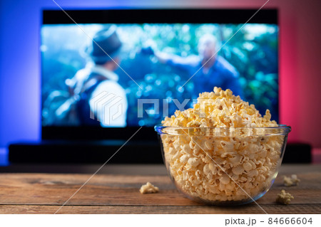 A glass bowl of popcorn and remote control in the background the TV works. Evening cozy watching a movie or TV series at home A glass bowl of popcorn and remote control in the background the TV works. Evening cozy watching a movie or TV series at home 84666604