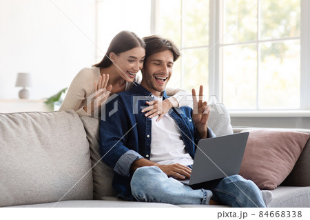 Smiling millennial european couple communicate with family remotely, wave hand, showing peace sign Smiling millennial european couple communicate with family remotely, wave hand, showing peace sign 84668338
