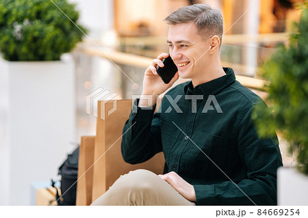 Portrait of handsome smiling young man talking on mobile phone sitting on bench in shopping mall looking away. Portrait of handsome smiling young man talking on mobile phone sitting on bench in shopping mall looking away. 84669254