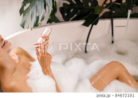 Relaxed young woman smiling while taking bubble bath and holding body care product, taking care of herself at luxury spa resort 84672101