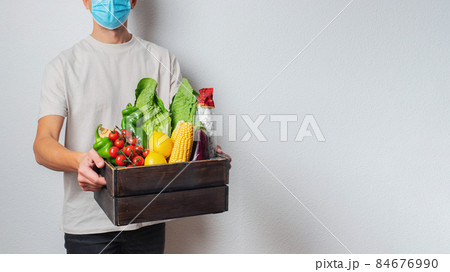 Young delivery man in protective face mask holding wooden box in hands. Man courier with shopping box. Safe online delivery from supermarket to home. COVID-19 coronavirus protection Young delivery man in protective face mask holding wooden box in hands. Man courier with shopping box. Safe online delivery from supermarket to home. COVID-19 coronavirus protection 84676990