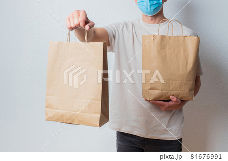 Young delivery man in protective face mask holding paper bags in hands. Man courier with shopping bag. Safe online delivery from supermarket to home. COVID-19 coronavirus protection 84676991