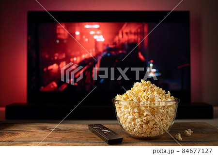 A glass bowl of popcorn and remote control in the background the TV works. Evening cozy watching a movie or TV series at home 84677127