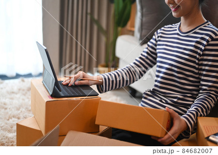 Smiling young woman confirming orders from customer on computer tablet and preparing parcel boxes for delivery. Smiling young woman confirming orders from customer on computer tablet and preparing parcel boxes for delivery. 84682007
