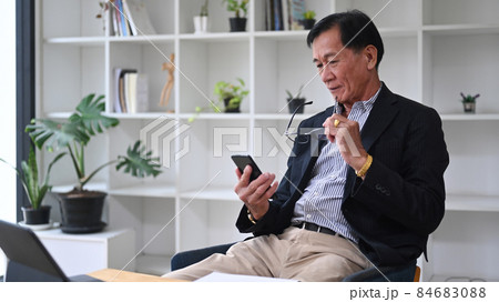 Mature businessman holding eyeglasses and using smart phone in office. 84683088