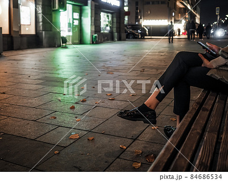 Beautiful elegant women's legs in black glossy loafers. A woman sits on a bench on a night street in the city in autumn 84686534