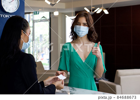 Caucasian tourist in green cloth  receiving a room card from hotel manager in black suit. Hotel staff and tourist wear face mask to protect them from the coronavirus outbreak. 84688673