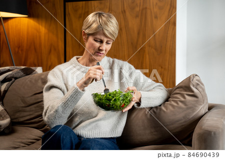 Sad middle-aged woman eats a tasteless green vegetable salad sitting on the couch Sad middle-aged woman eats a tasteless green vegetable salad sitting on the couch 84690439