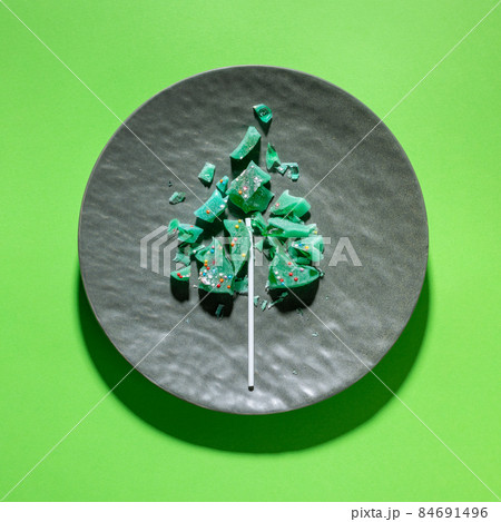 Creative sweet Christmas tree made of pieces of fractured green lollipop on textured grey plate on green. Trendy deconstruction. Festive joking foods image. Alternative Xmas tree. View from above. Creative sweet Christmas tree made of pieces of fractured green lollipop on textured grey plate on green. Trendy deconstruction. Festive joking foods image. Alternative Xmas tree. View from above. 84691496