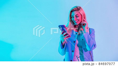 Happy fashionable young girl with a beauty smile in fashion elegant business clothes with a jacket holding a smart phone and typing a message on a multicolored neon pink light in the studio 84697077