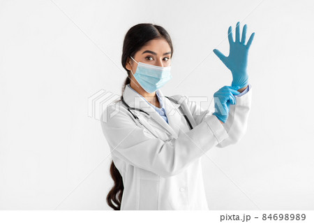 Serious millennial indian female doctor in coat and protective mask putting on hand rubber glove 84698989