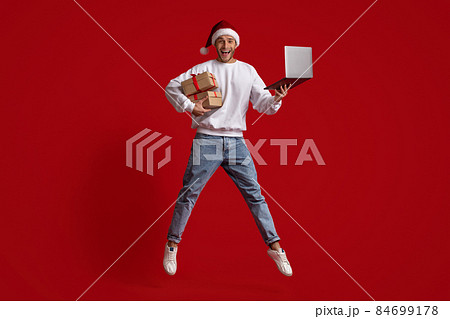Man In Santa Hat With Laptop And Gifts Jumping Over Red Background Man In Santa Hat With Laptop And Gifts Jumping Over Red Background 84699178