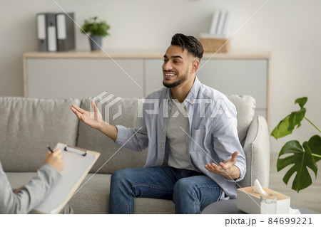 Young happy arab guy talking to his psychologist, receiving professional help, sitting on sofa at mental health clinic Young happy arab guy talking to his psychologist, receiving professional help, sitting on sofa at mental health clinic 84699221
