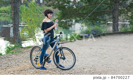 A brunette woman on a bicycle stopped by the river to drink water from a bottle 84700227