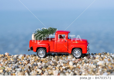 Red retro small car carrying christmas tree on the sea beach shore. Winter holiday concept 84703571