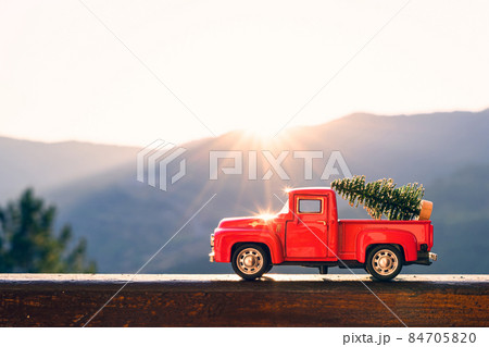 Red retro small car carrying christmas tree in the mountain in the sunbeams of sunrise. Winter holiday concept 84705820