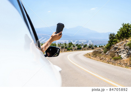 Woman driving a car holding paper cup of coffee from the open window. Trip on the serpentine road in the mountains. Summer vacation 84705892