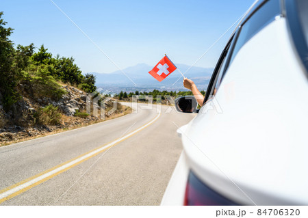 Woman holding Switzerland flag from the open car window driving along the serpentine road in the mountains. Concept Woman holding Switzerland flag from the open car window driving along the serpentine road in the mountains. Concept 84706320