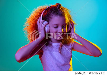 Portrait of little girl, kid listening to music in headphones isolated on blue studio background in neon light, filter. Portrait of little girl, kid listening to music in headphones isolated on blue studio background in neon light, filter. 84707713