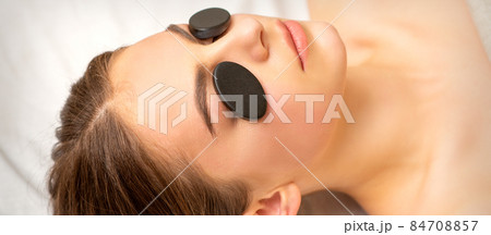 Black massage stones lying on the eyes of the young caucasian woman. Facial massage in a spa. 84708857