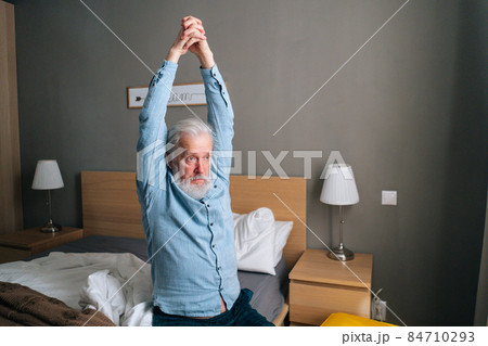 Senior adult man warming up and stretching hands up sitting on bed in morning after waking up at home. 84710293