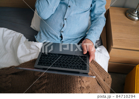 Close-up top view of unrecognizable mature adult male using typing on laptop computer sitting on bed with blanket at home in morning. 84710294