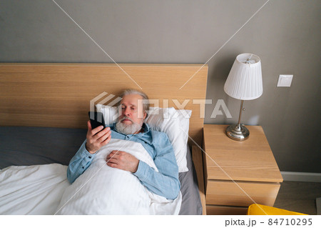 High-angle view of handsome bearded mature adult male lying on bed in hospital and talking on mobile phone by video call with family. 84710295