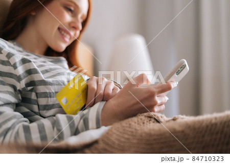 Close-up of smiling young woman input credit card details for online order on mobile phone lying on sofa at home. Close-up of smiling young woman input credit card details for online order on mobile phone lying on sofa at home. 84710323