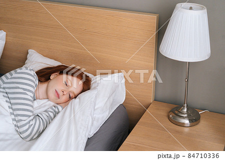 High-angle view of attractive redhead young woman sleeping well in bed hugging soft white blanket at home. High-angle view of attractive redhead young woman sleeping well in bed hugging soft white blanket at home. 84710336