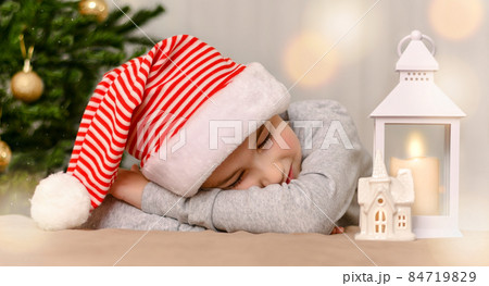 A child who fell asleep under the Christmas tree in a cap. With a happy smile and closed eyes, he lay down on hands. 84719829