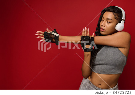 African American young woman with wireless headphones in gray sportswear stretches her arms against red background with copy space for ad African American young woman with wireless headphones in gray sportswear stretches her arms against red background with copy space for ad 84725722