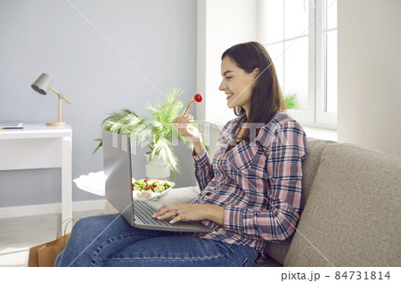 Happy woman using laptop computer while having lunch. Young girl sitting on couch at home, eating takeaway food, looking at PC screen, watching movie or browsing meal delivery website for discounts Happy woman using laptop computer while having lunch. Young girl sitting on couch at home, eating takeaway food, looking at PC screen, watching movie or browsing meal delivery website for discounts 84731814