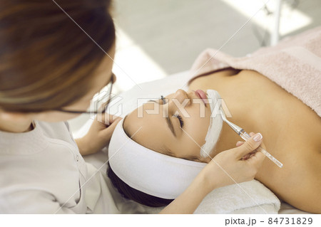 Professional cosmetologist at spa center applying anti acne kaolin clay mask on girl's face. Calm woman lying on bed in beauty parlor getting cosmetic facial treatment for fresh, beautiful, clear skin Professional cosmetologist at spa center applying anti acne kaolin clay mask on girl's face. Calm woman lying on bed in beauty parlor getting cosmetic facial treatment for fresh, beautiful, clear skin 84731829